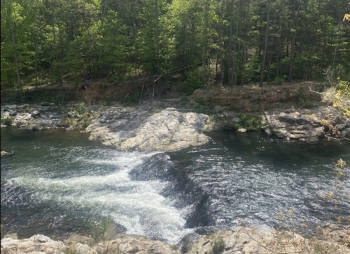 A serene river flows over rocks, surrounded by lush green trees in a peaceful natural setting.