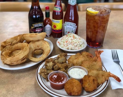 A plate of fried seafood, onion rings, coleslaw, and sauces, with drinks and condiments in the background.