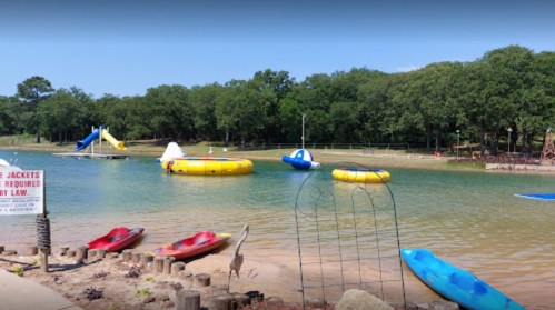 A serene lake scene with kayaks, inflatable slides, and a sign about life jackets, surrounded by trees.