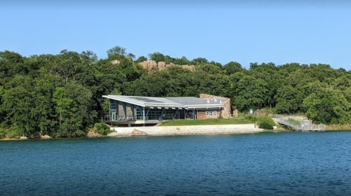 Modern building with a curved roof, surrounded by trees, by a calm lake under a clear blue sky.