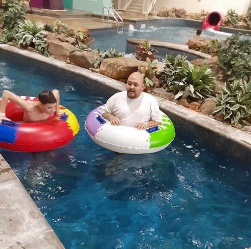 Two people float in a pool on colorful inner tubes, surrounded by rocks and greenery. A water slide is visible in the background.