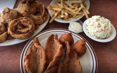 A plate of fried fish fillets, hushpuppies, coleslaw, onion rings, and French fries with a side of dipping sauce.