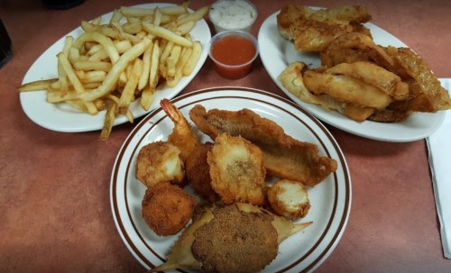 A spread of fried seafood, including fish fillets, shrimp, and hushpuppies, served with fries and dipping sauces.