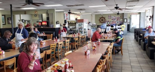 A busy diner with patrons eating at tables, staff serving, and a variety of food items on display.