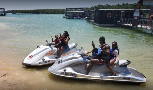 Two jet skis on a lake with four people, two on each, enjoying a sunny day and posing for the camera.