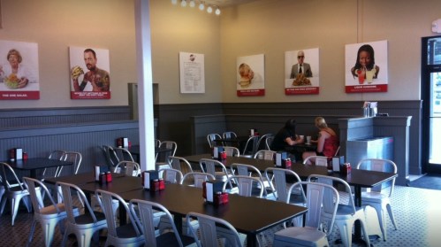 A spacious restaurant interior with metal chairs, tables, and food-themed wall art. Two patrons are seated at a table.