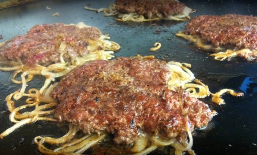 Four sizzling hamburgers cooking on a griddle with caramelized onions underneath.