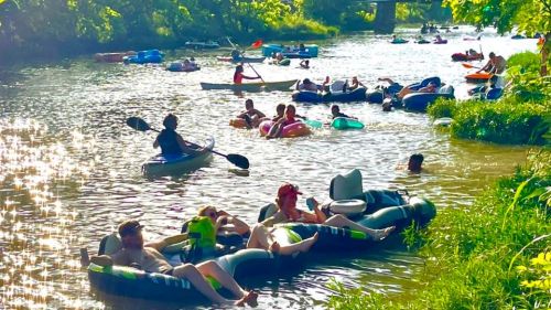 People relaxing on inner tubes and kayaks in a sunny river, surrounded by greenery and sparkling water.