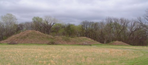 Two large earth mounds in a grassy field, surrounded by sparse trees under a cloudy sky.