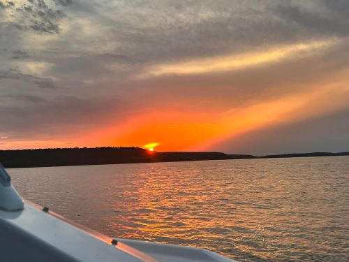 A vibrant sunset over calm waters, with orange and pink hues illuminating the sky and silhouetting distant hills.
