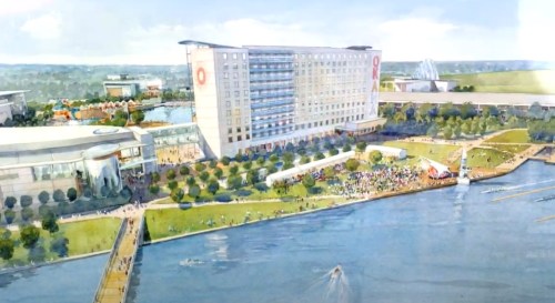 Aerial view of a waterfront development featuring a hotel, green spaces, and a crowd near the water.