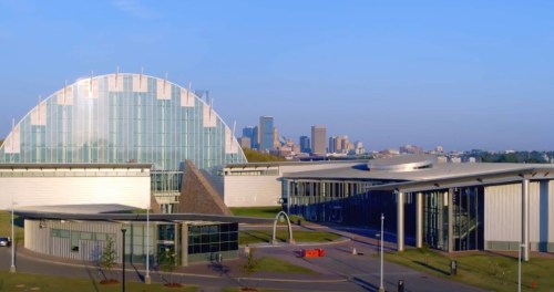 A modern architectural complex with glass structures and a city skyline in the background under a clear blue sky.