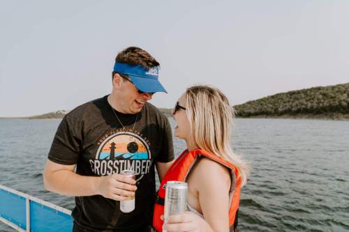 A couple smiles at each other on a boat, holding drinks, with a scenic water background. Both are wearing casual attire.