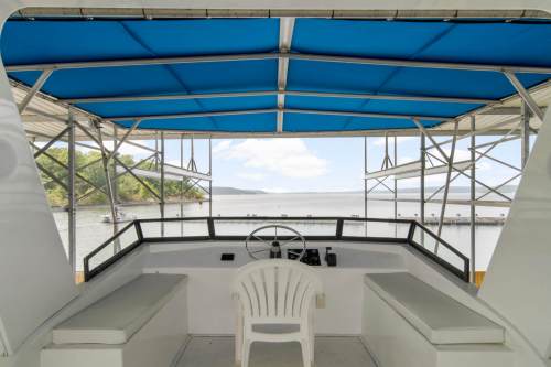 View from a boat's helm with a white chair, blue canopy, and a scenic waterfront in the background.