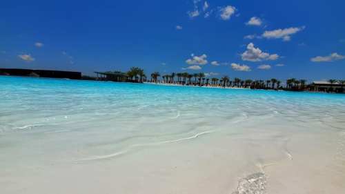 A serene beach scene with clear turquoise water, white sand, and palm trees under a bright blue sky with fluffy clouds.