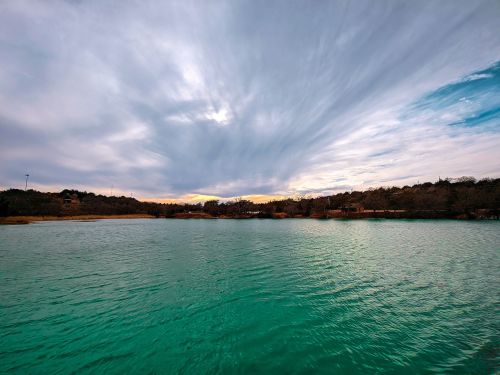 A tranquil lake with turquoise water under a cloudy sky at sunset, surrounded by trees and distant land.