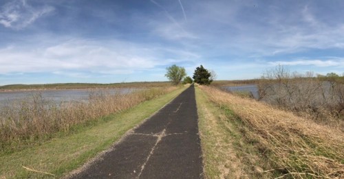 A paved path stretches between water bodies, bordered by grass and trees under a blue sky with wispy clouds.