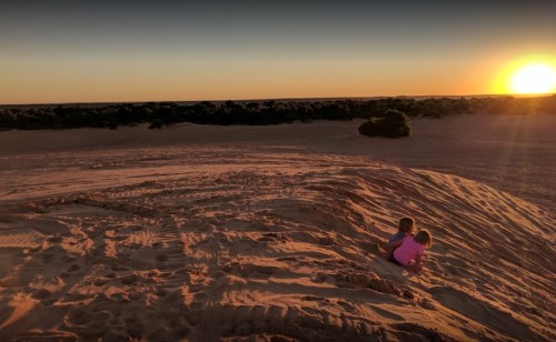 Two children sit on a sandy dune, watching a vibrant sunset over a desert landscape.