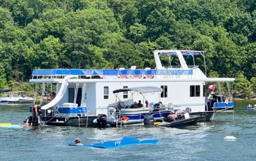 A houseboat on a lake surrounded by trees, with people swimming and enjoying the water.