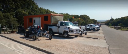 A parking area with various vehicles, including a truck and a trailer, surrounded by trees and a clear sky.