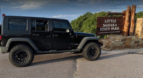 A black Jeep parked beside the entrance sign for Little Sahara State Park, surrounded by greenery.