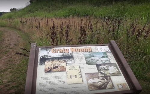 Sign for Craig Mound with historical information, surrounded by tall grass and a dirt path in a natural setting.