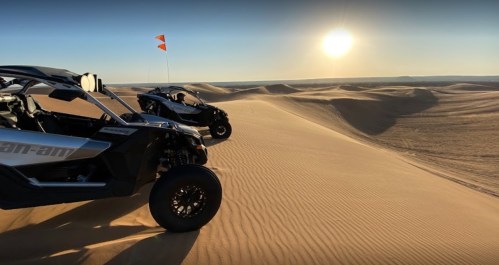 Two off-road vehicles parked on a sandy dune at sunset, with a clear sky and sun setting in the background.