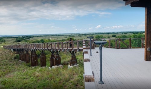 A wooden observation deck extends over a grassy landscape, with a person standing near the edge, overlooking the view.