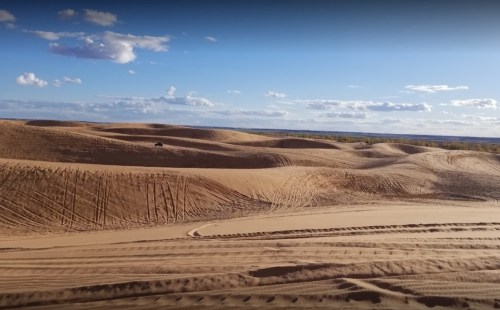 A vast desert landscape with rolling sand dunes under a blue sky and scattered clouds. Tire tracks are visible in the sand.