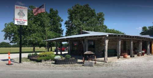 A rustic building with a porch, American flag, and sign for a local business, surrounded by greenery and gravel.