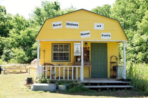 A small yellow building with a porch, labeled "Rural Oklahoma Museum of Poetry," surrounded by greenery.