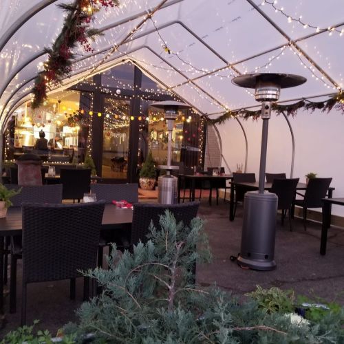 Cozy outdoor dining area with string lights, heaters, and festive decorations under a clear canopy.