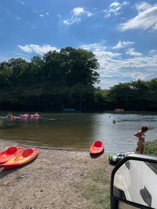 A sunny river scene with kayaks on the shore, people swimming, and lush trees in the background.