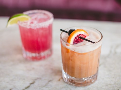 Two colorful cocktails on a marble surface: one with a lime wedge and the other garnished with an orange slice and cherry.