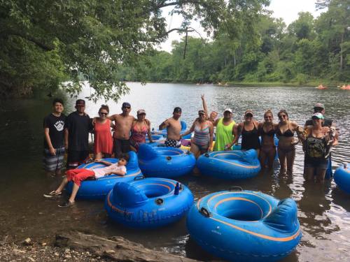A group of people in swimsuits stands by a river, surrounded by blue inner tubes, enjoying a sunny day outdoors.