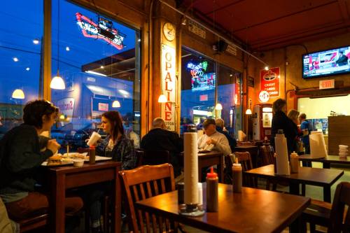 A cozy restaurant scene with diners enjoying meals, large windows showing a nighttime city view, and colorful decor.