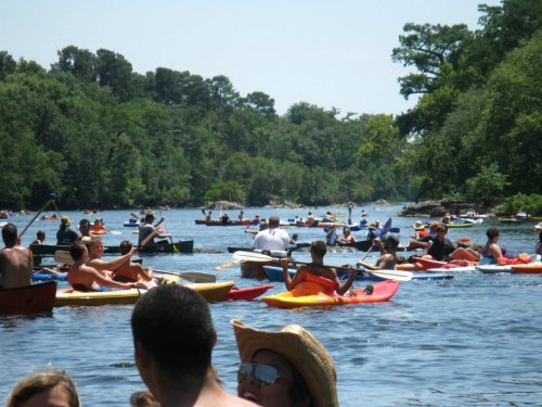 A busy river scene with many people kayaking and enjoying the outdoors on a sunny day. Lush greenery surrounds the water.