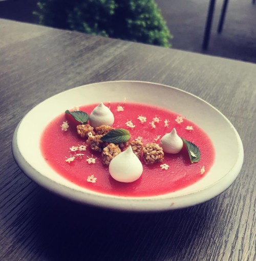 A vibrant pink soup garnished with meringue, granola, and mint leaves, served in a white bowl on a wooden table.