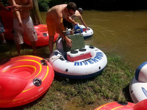 A group of people preparing colorful inner tubes by a river, with one person placing a cooler on a tube.