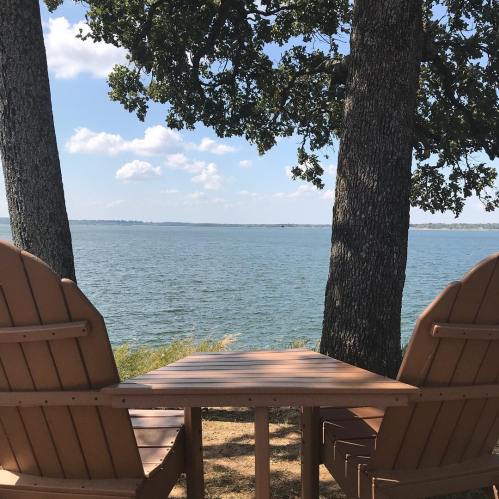 Two wooden chairs facing a serene lake, framed by trees under a blue sky with fluffy clouds.