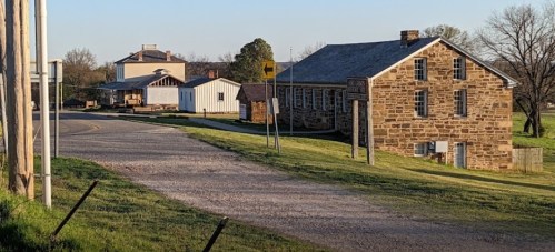A rural scene featuring a gravel road, stone buildings, and trees under a clear sky.