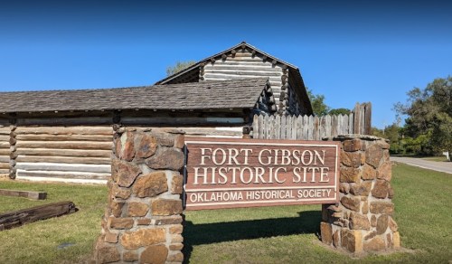 Sign for Fort Gibson Historic Site, featuring a log cabin structure and clear blue sky in the background.