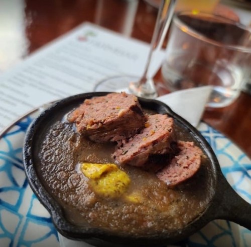 A small cast iron skillet with sliced meat on a bed of dark sauce, garnished with mustard, on a decorative plate.