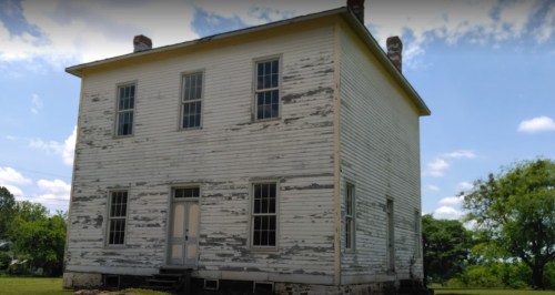 A weathered two-story house with peeling white paint and large windows, set against a blue sky and green trees.