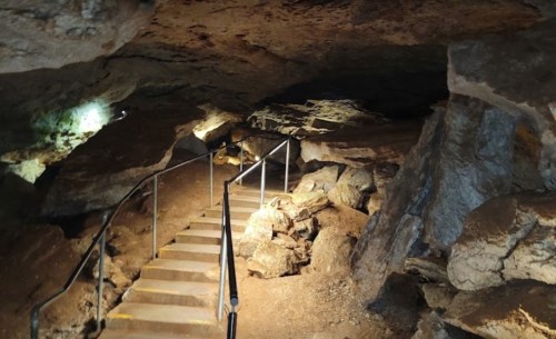A dimly lit cave interior with a staircase leading down, surrounded by rocky walls and scattered boulders.