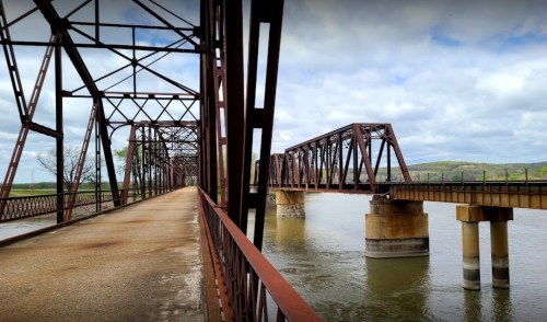Rusty metal bridge spans a river, with a pathway alongside and green hills in the background under a cloudy sky.