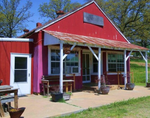 A rustic red building with a metal roof, featuring large windows and a porch, surrounded by greenery and potted plants.