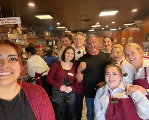 A group of smiling restaurant staff poses for a selfie with a man in a black shirt, inside a busy diner.
