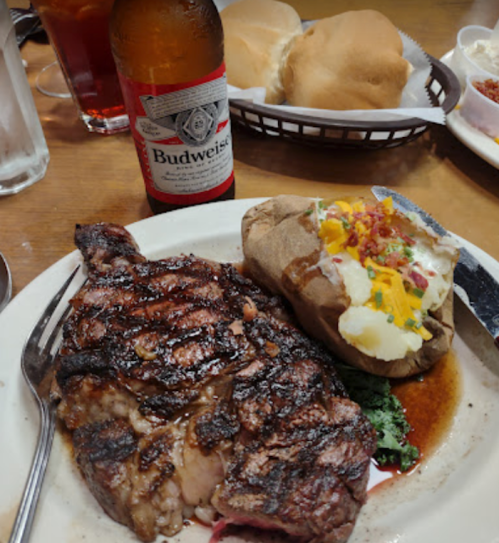 A plate with a grilled steak, baked potato topped with cheese and bacon, a Budweiser beer, and rolls in a basket.