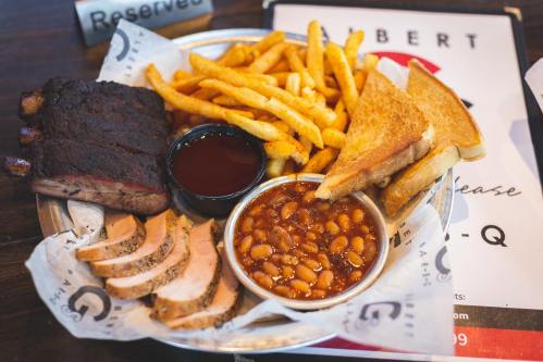 A platter featuring smoked ribs, sliced meat, fries, baked beans, and toasted bread, served with barbecue sauce.